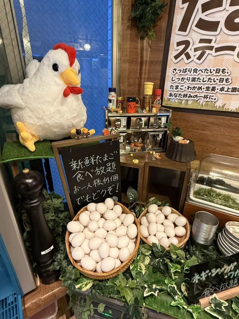 トッピングコーナー。新鮮なたまごの食べ放題サービスと薬味が並び、大きなニワトリのオブジェが飾られている。